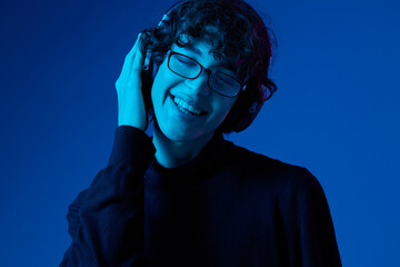 Teenage man wearing headphones listening to music and dancing and singing with glasses, hipster lifestyle, portrait blue background, neon light, style and trends, mixed light, copy space