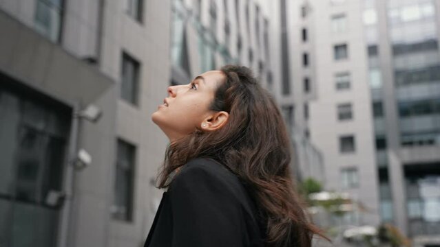 Young Caucasian female with dark long hair walking in city center around grey skyscrapers, girl likes view of high contemporary architecture.