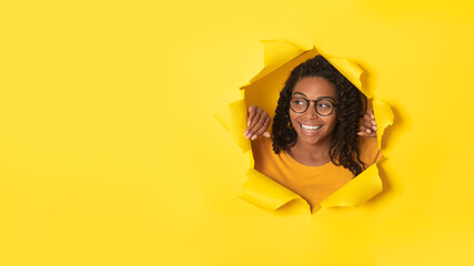 Excited african american lady in glasses posing in torn paper, looking at free space through hole on yellow background © Prostock-studio