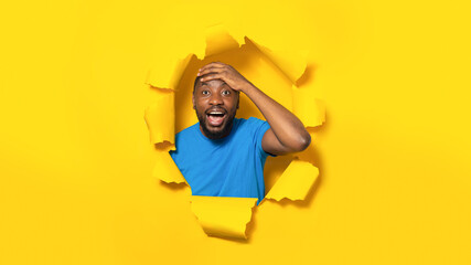 Portrait of overjoyed black man touching head and breaking through hole in yellow paper background,...
