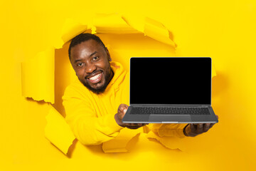 Excited black man holding laptop with empty blank screen showing device through yellow paper torn hole, mockup