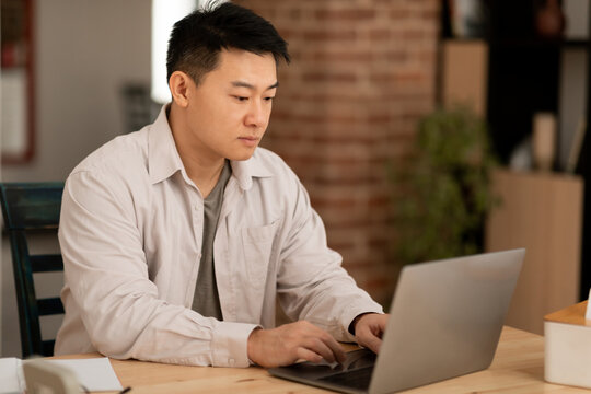 Serious middle aged asian man using laptop computer, typing on keyboard while working online, sitting at home office