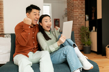 Overjoyed japanese couple sitting on couch with digital tablet, celebrating online win at home, copy space
