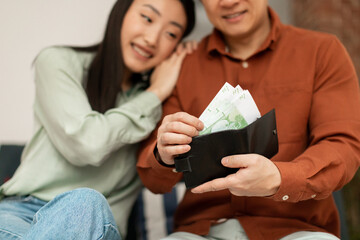 Money saving concept. Happy korean couple putting banknotes into wallet, sitting on sofa at home, selective focus