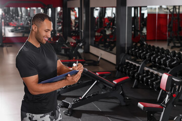 Fototapeta na wymiar Happy trainer writing down workout plan in modern gym, space for text