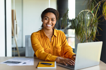 Portrait of happy black businesswoman working on laptop and smiling at camera, sitting in office interior, copy space