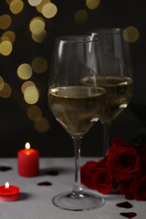 Glasses of white wine, rose flowers and burning candles on grey table against blurred background. Romantic atmosphere