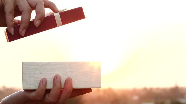 Close-up of a happy woman opening a gift box with a white ribbon and a surprise gift over a sunset or the sun in a natural outdoor background. Valentine's Day, a gift box, a gift, Xmas, Christmas, 4K
