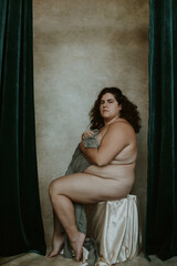 portrait of a plus size woman with curly hair sitting on a stool looking at camera clutching gray fabric