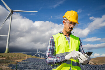An engineer in a field of wind turbines and solar panels works using his phone, renewable energy
