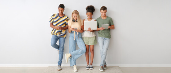 Group of teenagers with different devices standing near light wall