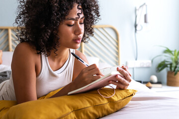 Pensive young multiracial latina woman lying down on bed with serious expression writing on journal...