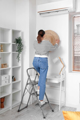 Young woman on stepladder hanging rattan pouf onto wall at home