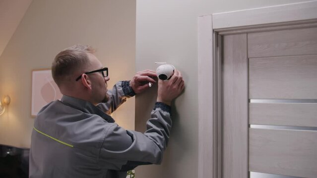 Installer in uniform puts security camera on wall fastening and connects it to system with cable. Man installs cameras in house. Concept of CCTV cameras, monitoring, safety and privacy. Zoom in shot.