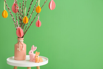 Vase with willow branches and Easter rabbits on table near green wall
