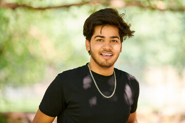 Smiling young indian man candid portrait in black t shirt and silver neck chain on outdoor public...