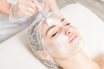 Beautician applies vitamins cream mask on woman face for rehydrate face skin, anti aging cosmetic procedure in beauty spa salon. Cosmetologist in gloves holds cosmetic brush for applying mask