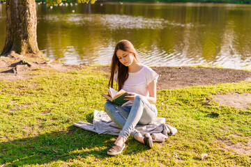 Young hipster girl enjoys sun and nice warm day reading book while relaxing, woman is relaxing outdoors sitting next to a lake in a park in summer, a caucasian female student in the sun