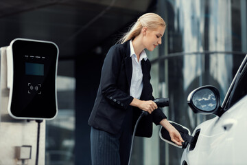 Progressive businesswoman insert charger plug from charging station to her electric vehicle with...