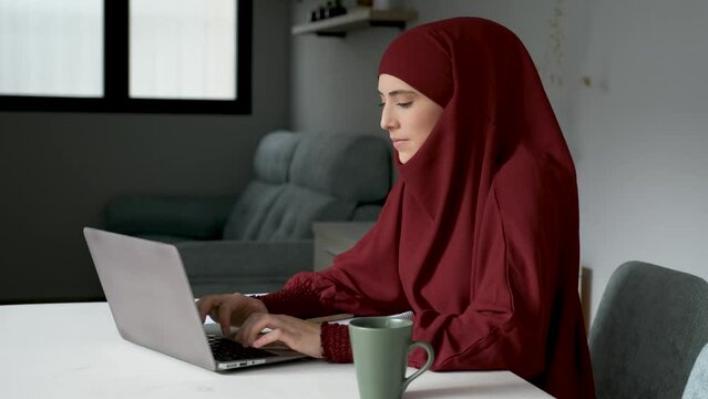 Muslim young woman in hijab on a phone call while working in a laptop at home.