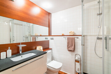 Bathroom with one wall of deep caramel wood paneling and large mirror between them. In contrast, white sink is built into black marble countertop. Mirror reflects shower cabin with glass doors.