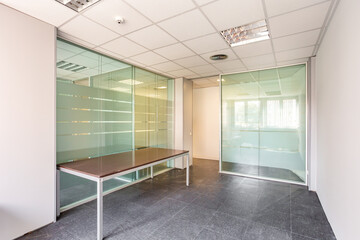 In a bankrupt office building, one of the many empty rooms with transparent, solid glass wall panels and an outdated ceiling covering is in need of repair and refurbishment.