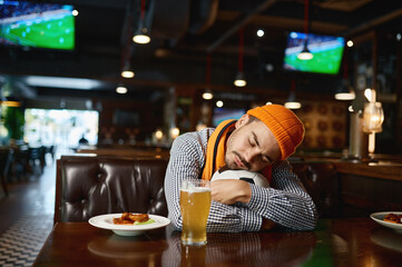Funny young man soccer fan sleeping on ball while rest in sport bar