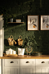 Kitchen interior with wicker baskets and paintings in wooden racks on a green wall, cutlery, dishes, towels and plants on a wooden table. Kitchen in warm sunlight.