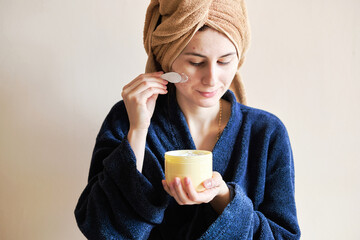 woman in a robe and with a towel on her head applies cosmetics to skin