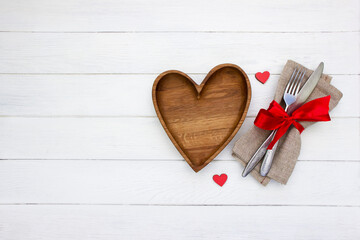 Valentine's Day place setting. Wooden heart shape plate, fork and knife with red bow, red ribbon on white wooden board