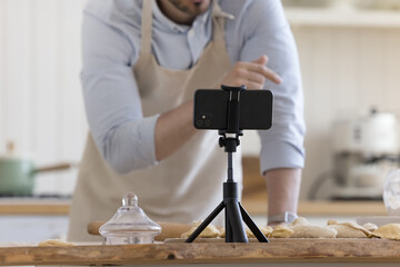 Male baker preparing pastry food, speaking at smartphone webcam placed on kitchen table. Cook...