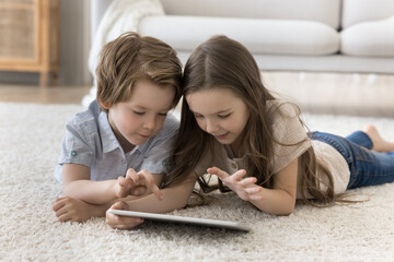 Curious little brother and sister kids using tablet for Internet communication, lying on warm soft carpeted floor, playing video game, browsing blogs, chatting, enjoying leisure alone