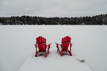 Red adirondack chairs on end of snowy dock on a lake in winter.