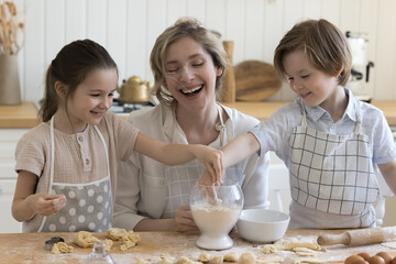 Joyful young mother and two happy pretty little kids in aprons baking together in home kitchen, having fun, laughing at floury smudges on faces, shaping dumplings, cookies from dough at table