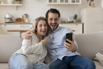 Fototapeta premium Cheerful positive young dating couple in love posing for selfie, taking self video, hugging on home sofa, showing Victory fingers gesture at smartphone, smiling, laughing, having fun