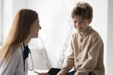 Joyful kid visiting pediatrician, sitting on medical couch in office, smiling, laughing. Happy family doctor woman asking little patient about health complaints, examining boy