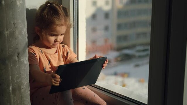 Child girl age 2 years old uses a tablet while sitting on the windowsill at home.Education at home. City life. Sunny morning.