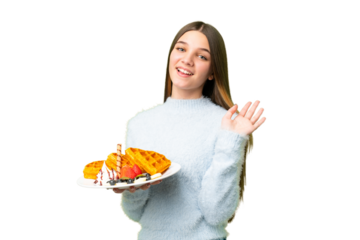 Teenager girl holding waffles over isolated chroma key background saluting with hand with happy expression