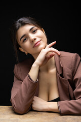 Fototapeta premium Classic studio portrait of a young brunette dressed in a black top and formal suit, who is sitting on a chair against a black background.