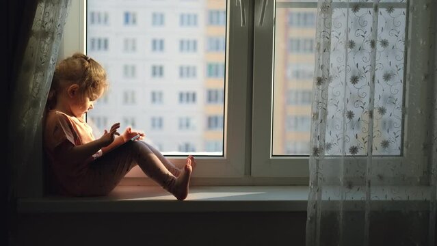 Child girl age 2 years old uses a tablet while sitting on the windowsill at home.Education at home. City life. Sunny morning.