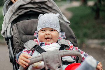Baby boy in warm colorful knitted jacket sitting in modern stroller on a walk in a park. Child in...