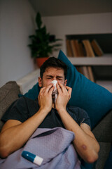 A young man blowing his nose and feeling virus symptoms. An ill young man lying down at home under a blanket and blowing his nose.