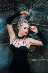 A slender beautiful woman with blond hair in a black vintage dress and expensive jewelry, stylish makeup and hairstyle, stands near the walls of an old castle.