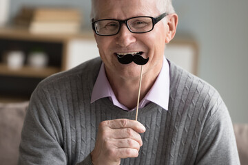 Happy positive senior man applying fake paper moustache on stick to face, celebrating movember,...