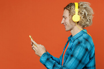 Side view smiling young blond caucasian man wear blue shirt orange t-shirt headphones listen to music use mobile cell phone isolated on plain red background studio portrait. People lifestyle concept.