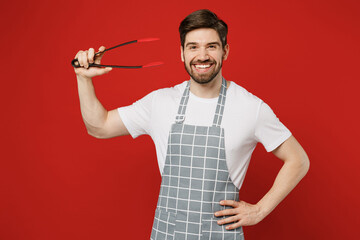 Young fun housewife housekeeper chef cook baker man wears grey apron hold in hand kitchen salad serving plastic pair of tongs for grill isolated on plain red background studio. Cooking food concept.