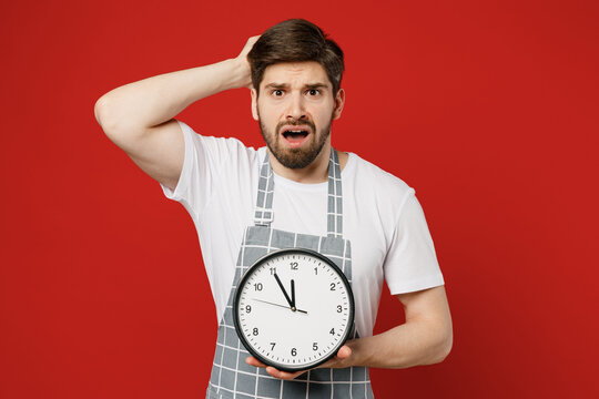 Young confused scared shocked displeased disappointed male housewife housekeeper chef cook baker man wear grey apron hold head show clock isolated on plain red background studio. Cooking food concept.