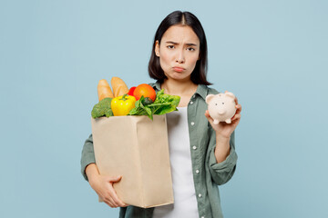 Fototapeta na wymiar Young sad displeased woman wear casual clothes hold brown paper bag with food products pig money box isolated on plain blue cyan background studio portrait. Delivery service from shop or restaurant.