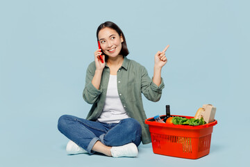 Full body fun young woman in casual clothes hold red basket with food products talk speak on mobile...