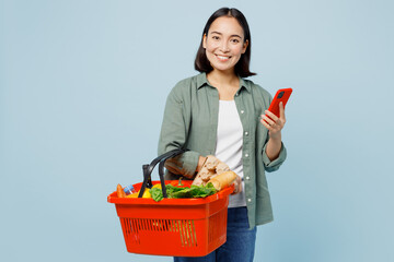 Plakat Young satisfied happy fun woman in casual clothes hold red basket with food products use mobile cell phone isolated on plain blue background studio portrait. Delivery service from shop or restaurant
